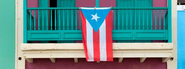 The Puerto Rican flag, with a white star inside a blue triangle backed by five red and white stripes, hangs over the balcony of a colorful old building.