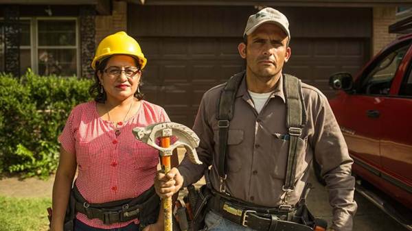 Two workers stand looking at the camera. A woman wearing a yellow hard hat and a man holding a building tool