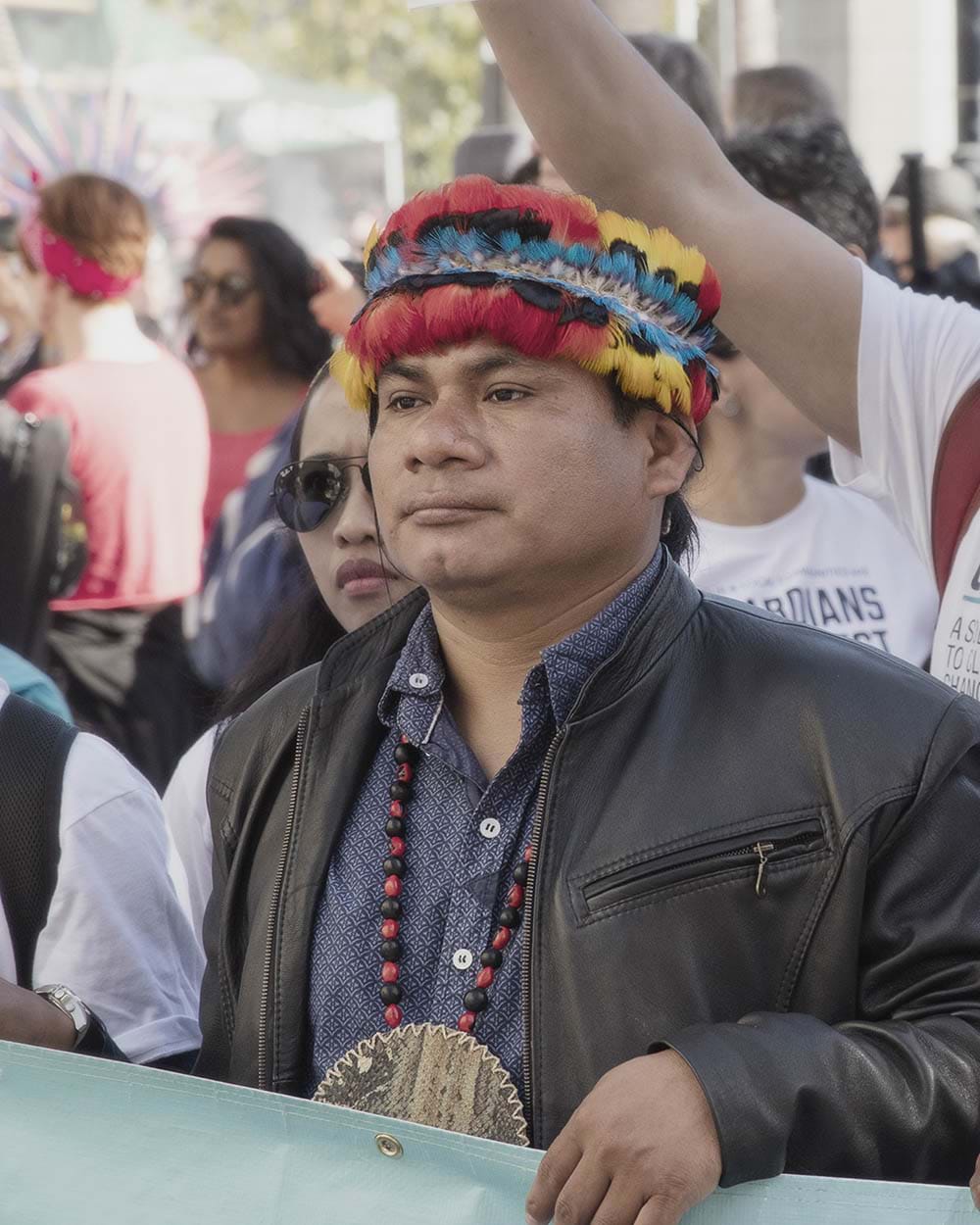 An Indigenous man with feather headdress and wearing a leather jacket and a blue button down t-shirt holding a sign at a street protest. 