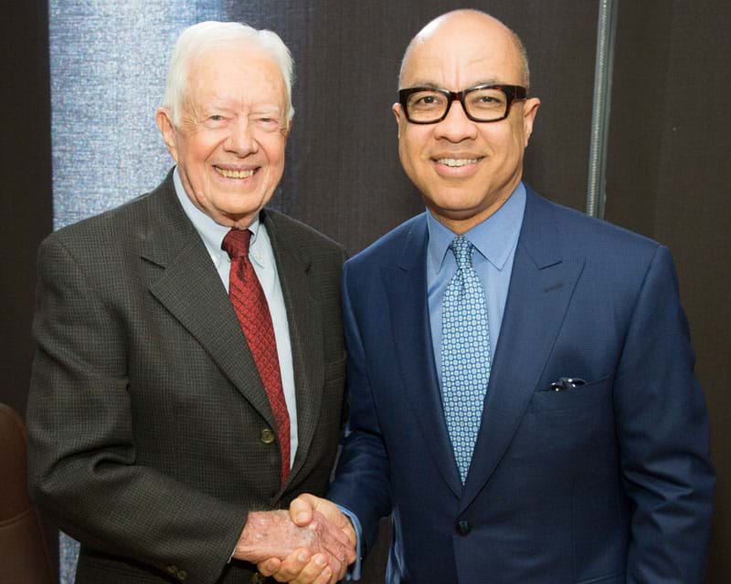 President Jimmy Carter shaking Darren Walker’s hand at the Ford Foundation. Both are smiling and wearing suits.