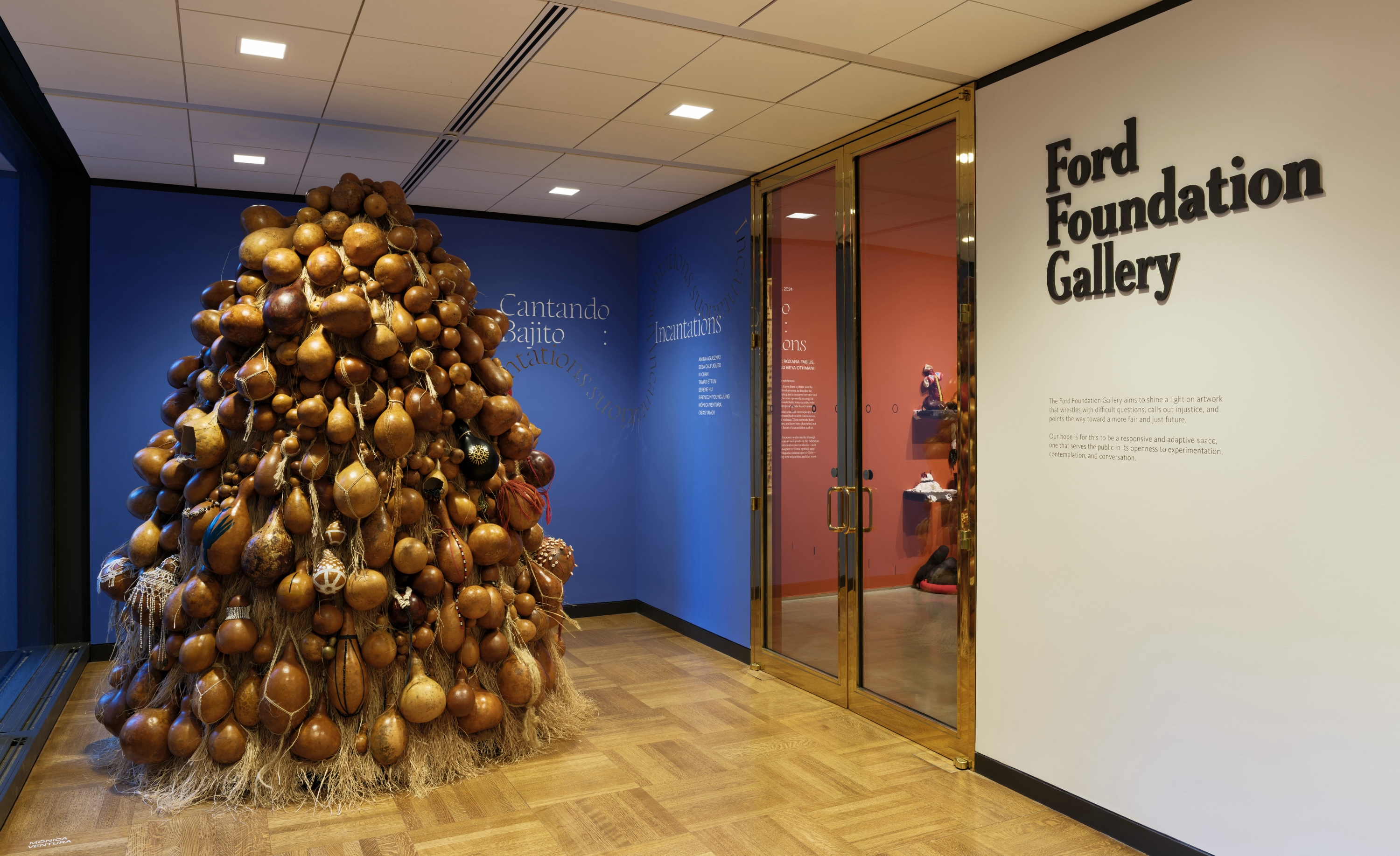A sculptural artwork before the entrance of the Ford Foundation Gallery that stands before a blue painted wall and on top of a wood floor. The sculpture is made up of piled gourds and straw, and stands 8 feet tall.