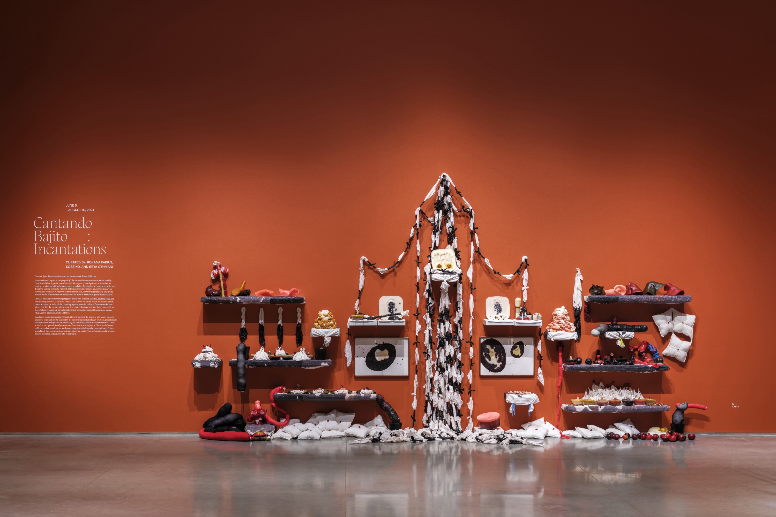 A terracotta-red wall connecting to a gray floor. On the left side of the wall is exhibition text with the title “Cantando Bajito: Incantations”. To the middle and right of the wall is a mixed media installation resembling a shrine with soft sculpture, pillows, fabric chains, and wax molds.