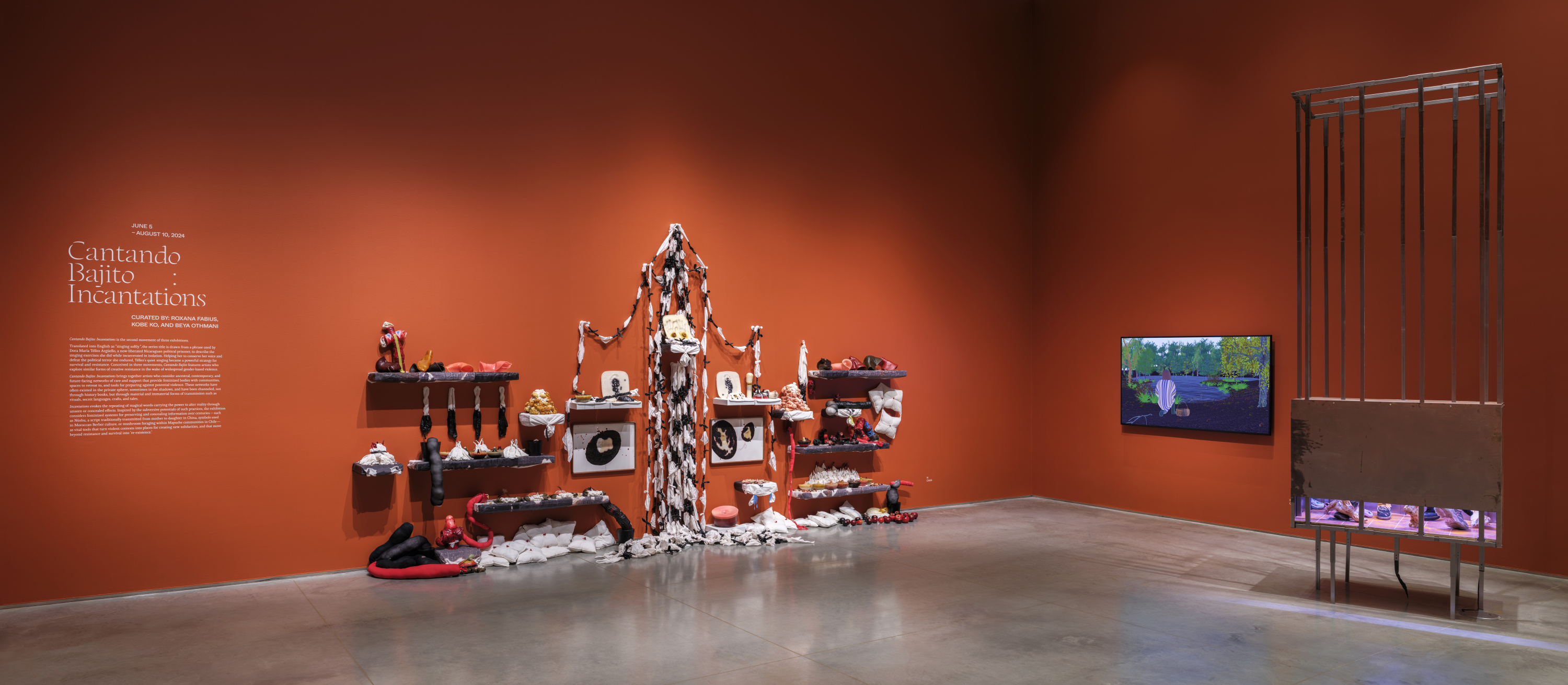 Gallery interior with terracotta-red walls and a gray floor. There are several artworks displayed - from left to right - a soft-sculpture mixed-media installation, a tv monitor with a video animation, and a ceiling-height cage sculptural installation.