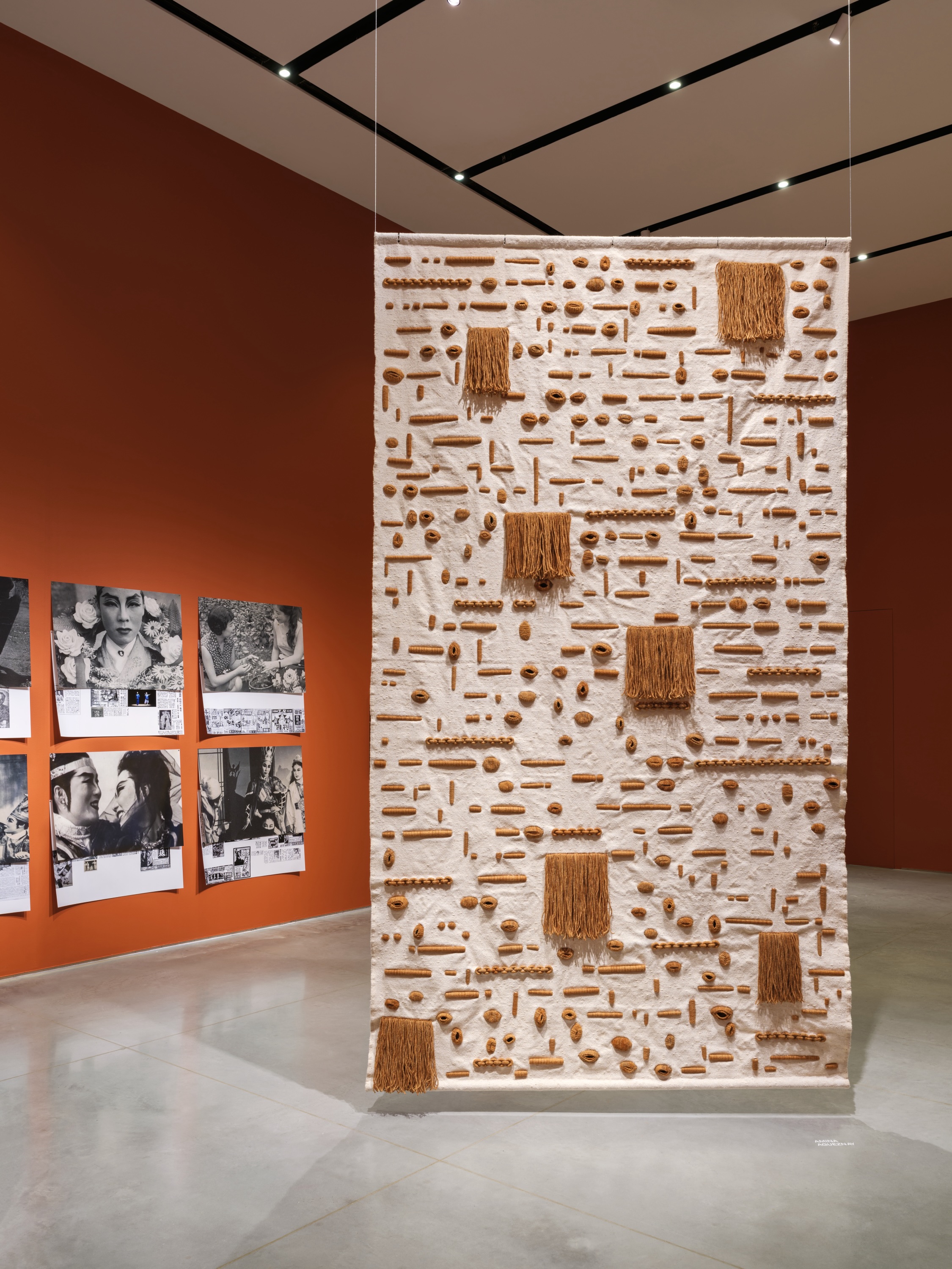 Gallery interior with terracotta-red walls and a gray floor. On the left wall is a photo collage installed in a grid featuring historical photographs of East Asian stage performers and Korean newspaper cuttings. Suspended from the ceiling in the middle of the gallery floor is a sand-colored tapestry with brown geometric details.