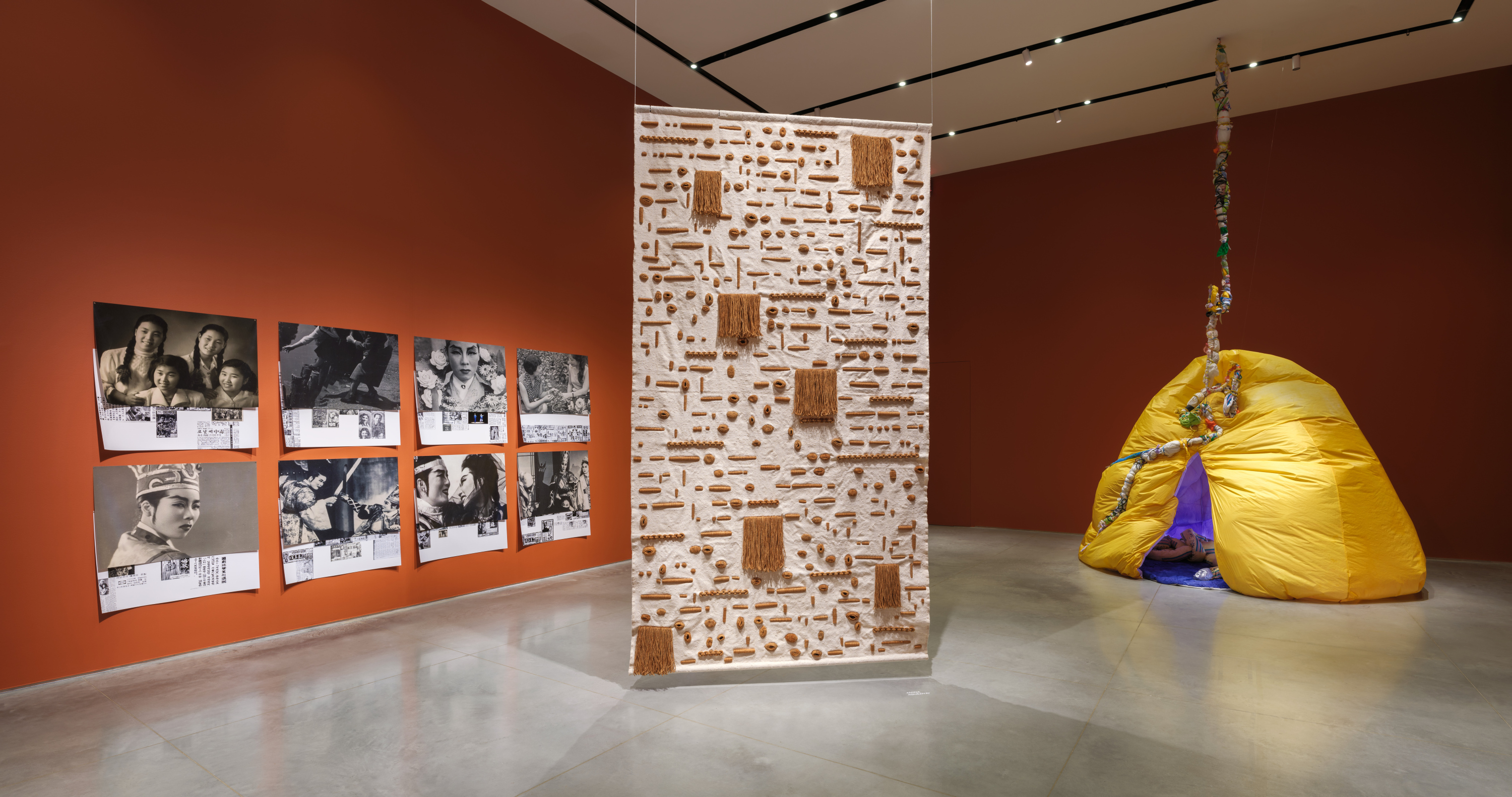 Gallery interior with terracotta-red walls and a gray floor. On the left wall is a photo collage installed in a grid featuring historical photographs of East Asian stage performers and Korean newspaper cuttings. Suspended from the ceiling in the middle of the gallery floor is a sand-colored tapestry with brown geometric details. To the right is an inflatable yellow hut, split vertically with a blue and purple plush interior. A fabric snake is coiled on the floor.