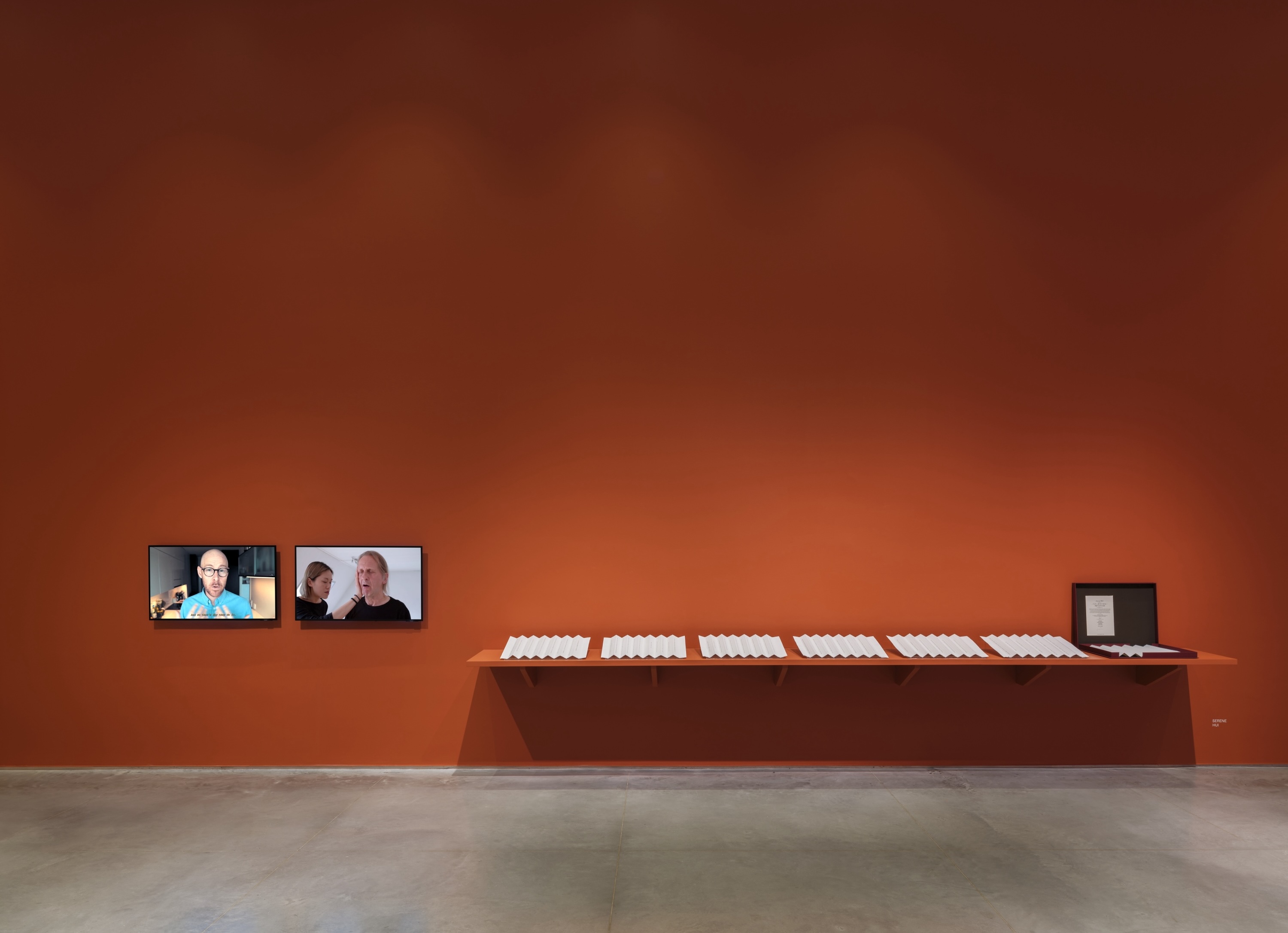 A terracotta-red wall connecting to a gray floor. On the left are two monitors displaying a left and right-channel video. The left-channel video alternates between shots of mouts speaking and footage from popular culture films and television. The right-channel video features a white man and an East Asian woman practicing vocal exercises. To the middle and right, is a terracotta-red shelf holding a row of seven sheets of embossed paper folded accordion-style. The last paper on the end-right is placed in a red box.