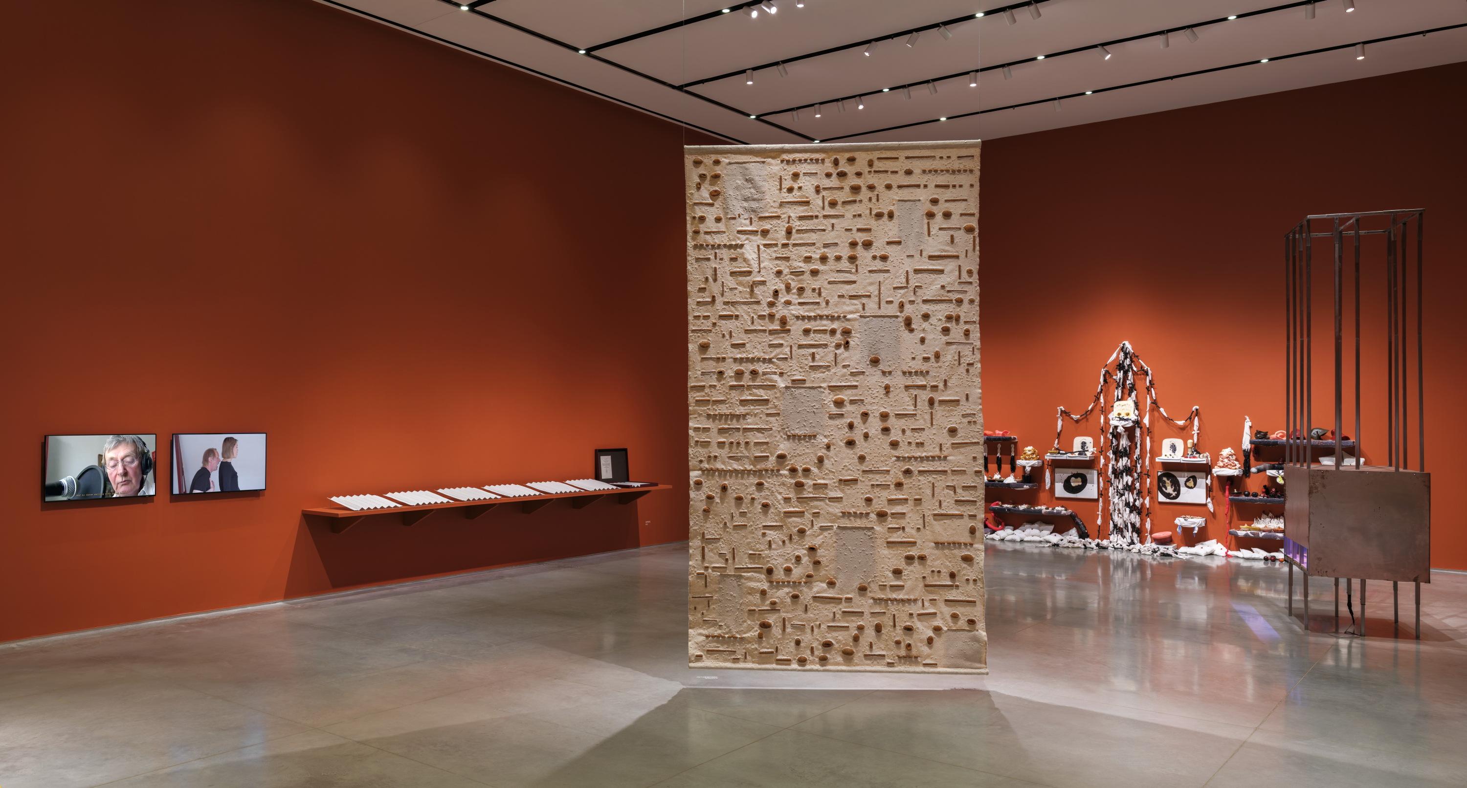 Gallery interior with terracotta-red walls and a gray floor. There are artworks installed on the wall, two tv monitors, a suspended tapestry, and a free-standing cage sculpture.