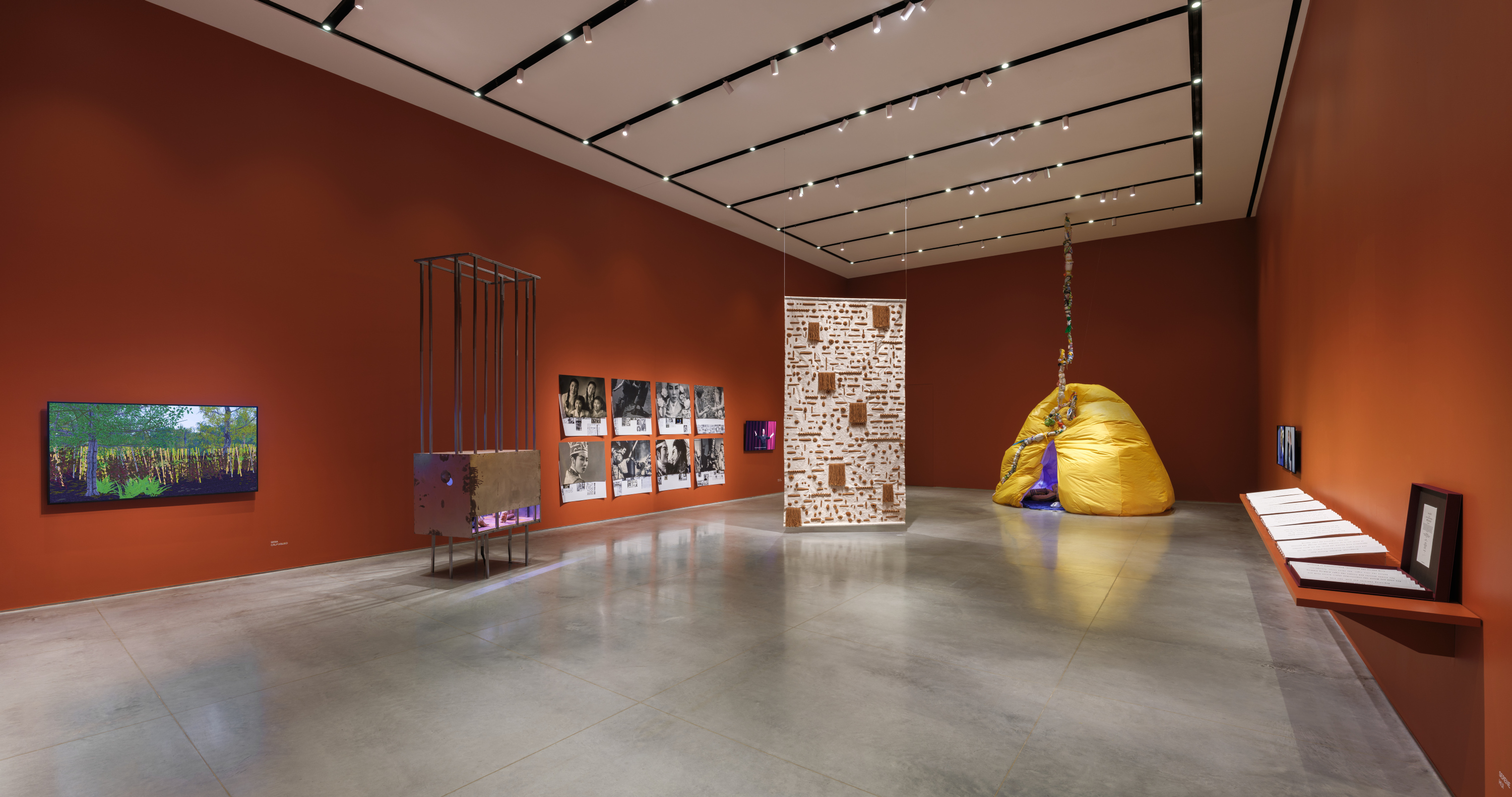 Gallery interior with terracotta-red walls and a gray floor. There are artworks installed on the wall, tv monitors, a suspended tapestry, a free-standing cage sculpture, and an inflatable yellow hut.