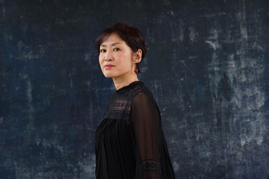 Dr. Akiko Mizoguchu with short dark hair is wearing a black outfit, standing against a dark, textured background, looking slightly to her right.