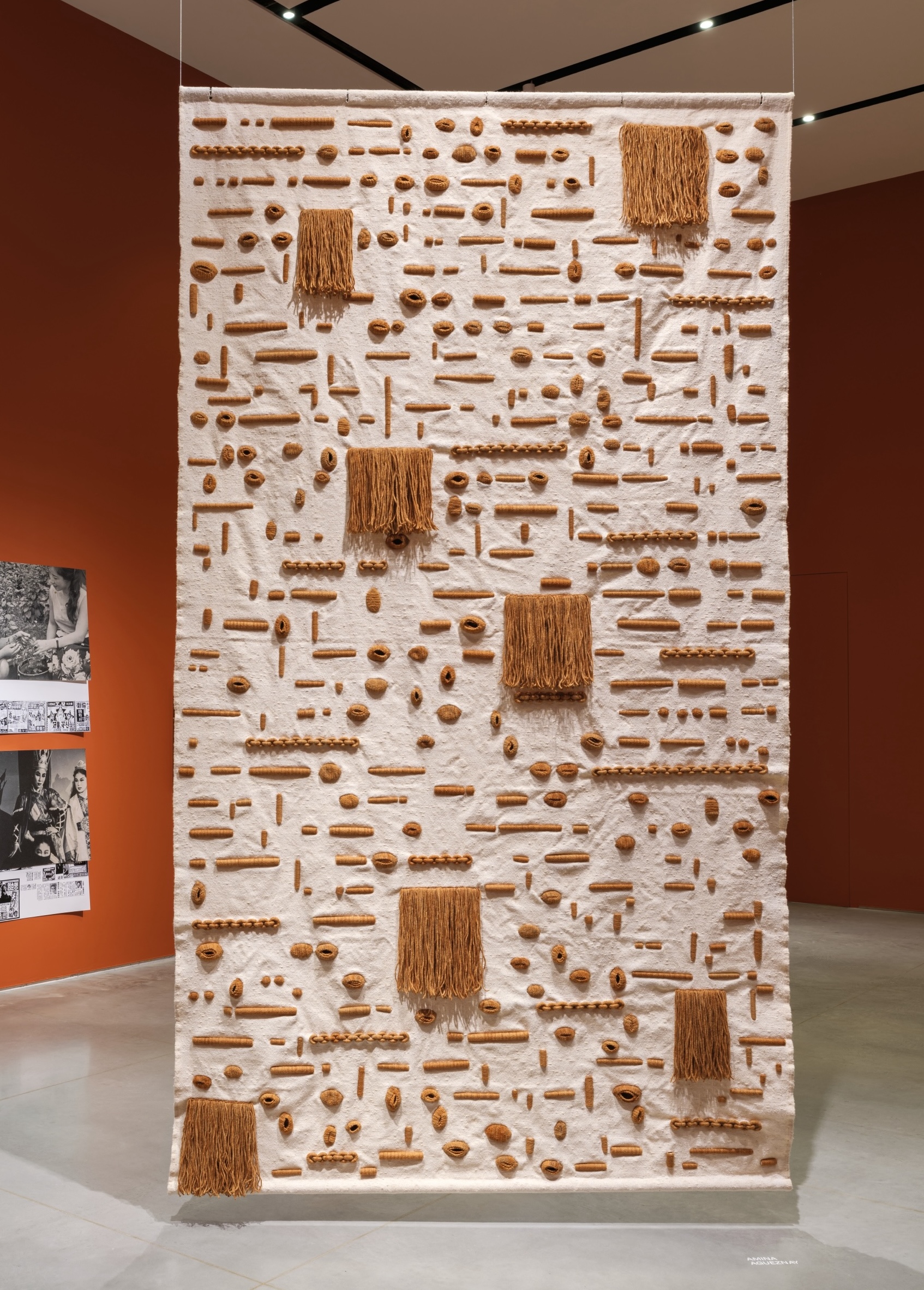 Photo of a sand-colored tapestry with brown geometric details suspended from the ceiling of a gallery.