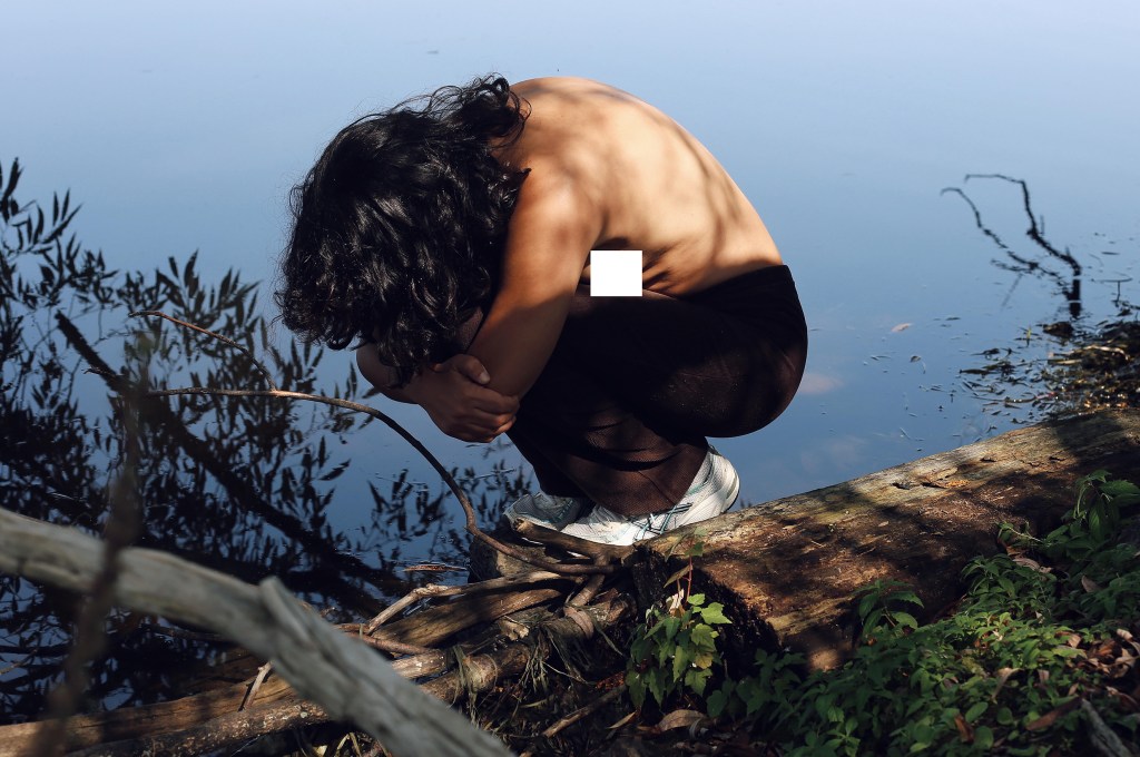 A person with long hair, bare back, and dark pants sits curled up on a log by a tranquil body of water. They are hugging their knees with their arms and resting their head on their knees. The surrounding area has branches, leaves, and reflections in the water.