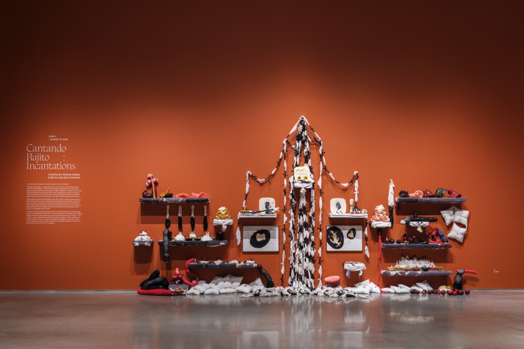 Gallery interior with terracotta red walls featuring exhibition text to the left and mixed media installation resembling a shrine with soft sculpture, pillows, fabric chains, and wax molds to the right.