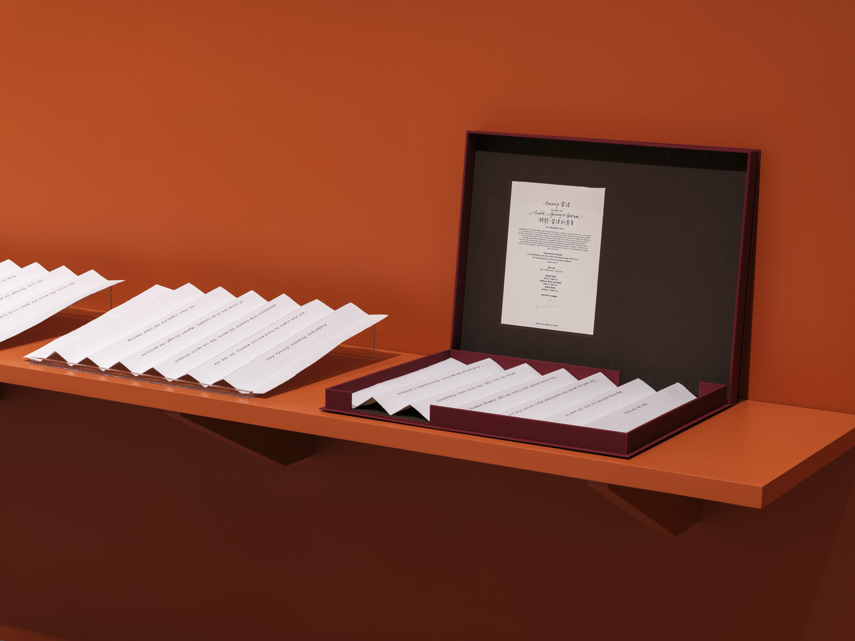 A shelf displaying sheets of thick embossed paper folded accordion-style, where each fold contains a line of text. The last paper to the right is displayed lying down in an opened dark red box. The cover of the dark red box is on its side displaying a certificate.