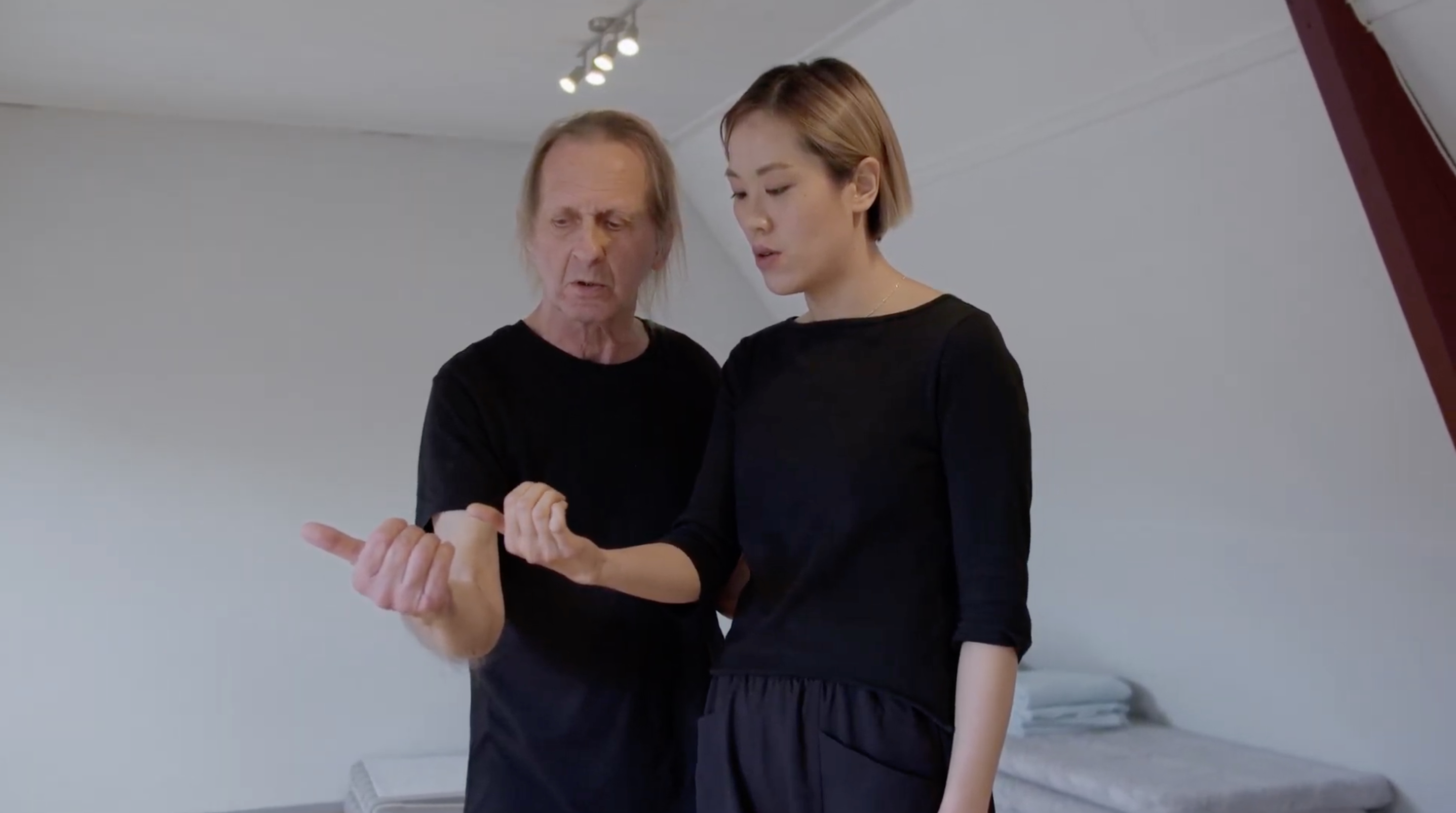 A screenshot of a 12:44 video of a white man and an East Asian woman practicing vocal exercises. They both are staring at their hands, and lifting one hand up in a fist and thumbs-up motion.