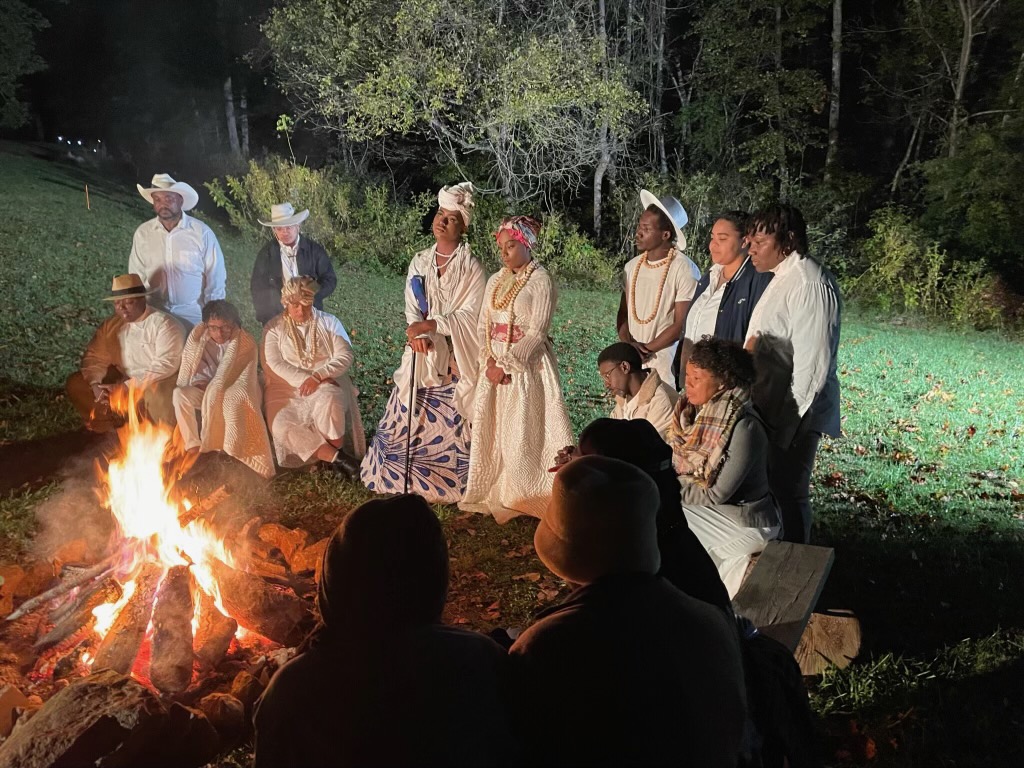A group of people is gathered around a bonfire at night. Some individuals are dressed in colorful traditional attire with hats and headscarves, while others are in casual clothing. The background features trees and grass, illuminated by the fire's light.