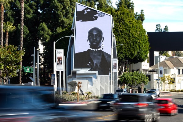 A large black and white billboard features a portrait of an individual wearing jewelry and a dark outfit. The billboard is set against a backdrop of tall trees and buildings while cars drive by in the foreground on a busy street. Artwork by Chris E. Vargas.