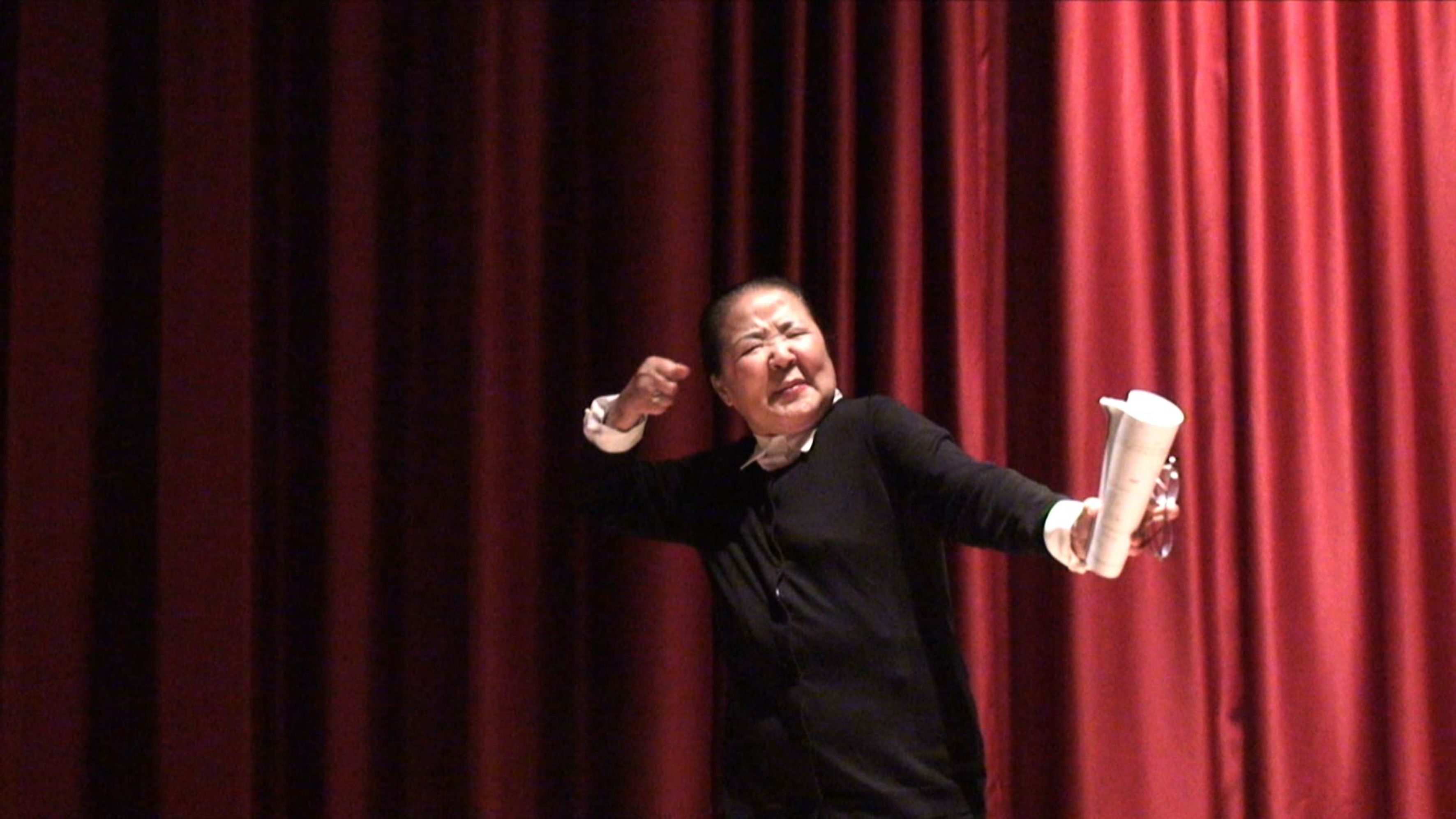 A still from a 10:14 video of an East Asian woman on a stage giving directing notes in Korean in front of a red curtain backdrop.