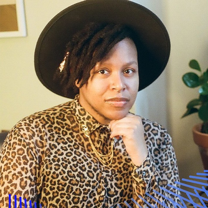 Cyrée, a Black trans person in a leopard print shirt and wide-brimmed black hat, rests his head on his hand. Behind him are a potted plant and a tarot card on a glass table.