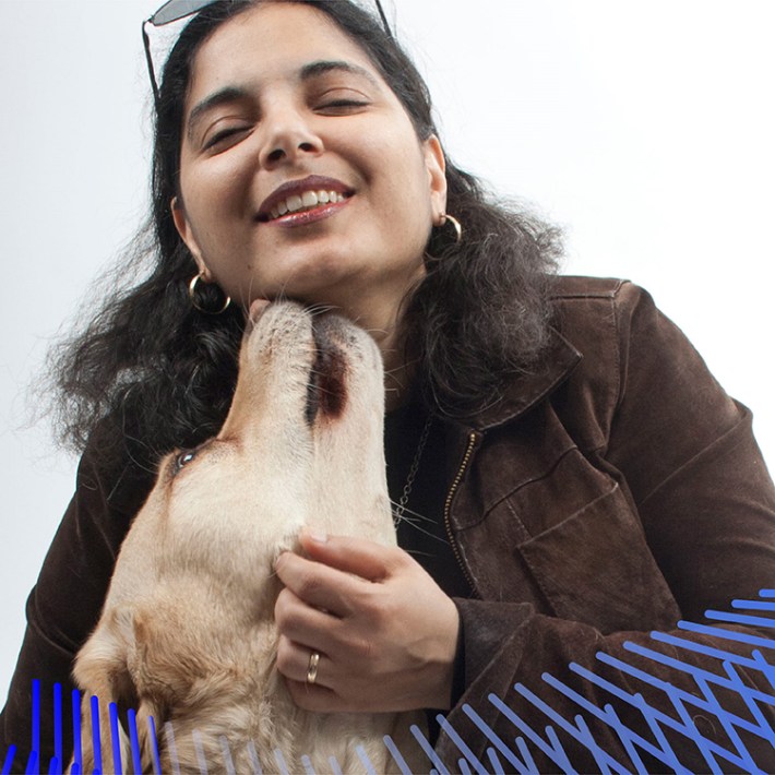 Day, an Arab-American woman in a suede jacket with brown skin and dark hair, sits with her eyes closed. In front of her, a yellow lab guide dog looks up and licks under her chin.