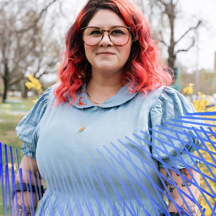 Emily, a white woman with electric pinkish orange long loose curly hair, oversized tortoiseshell glasses, and a light blue dress with an embroidered red scorpion on the collar. She has crutches and various tattoos all over her arms, and she stands in a park on a sunny day with yellow flowers behind her. She is smiling and looking directly at the camera.