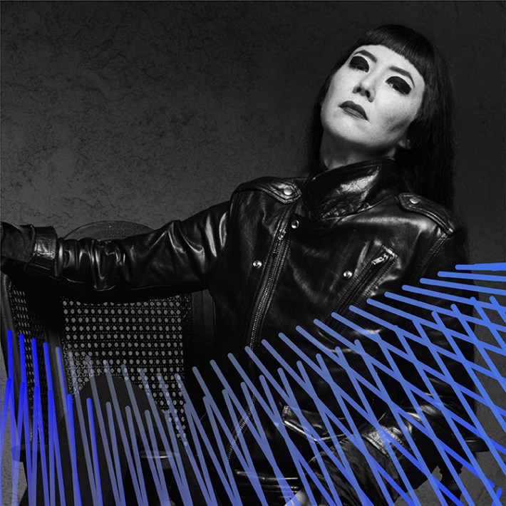 Johanna, a Korean American femme, wears a black leather jacket, black pants, and black platform Rick Owens boots. They sit in a wooden chair with one leg propped up on the arm rest.