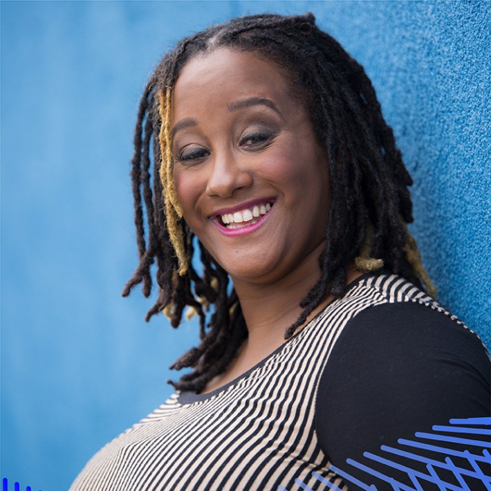 Kayla poses in profile in front of a light blue background. She is a milk chocolate-skinned Black woman who is wearing a long sleeve black and tan striped shirt. She has shoulder length dreadlocks and a glowing smile.
