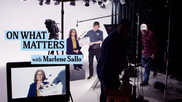 A behind-the-scenes view of a TV studio setup. Marlene Sallo sits in front of the camera, holding a clapperboard with "On What Matters with Marlene Sallo" displayed beside her. Crew members adjust equipment and lighting. A monitor shows her on-screen image.