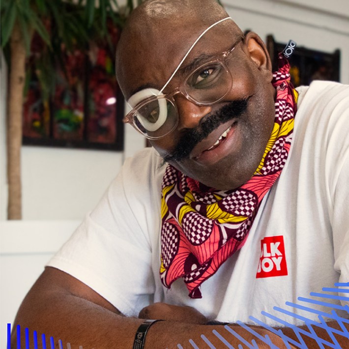 Saleem, a Black man with a partially paralyzed face, an ever-widening smile, and a handlebar mustache, faces the camera. He wears clear-framed glasses, a plastic moisture chamber over his right eye, and a right-sided cochlear implant and sits comfortably hunched over a table with arms loosely folded. He wears a white T-shirt with “Blk joy” printed inside a red square and a pink and yellow floral-print bandana around his neck. Two pamphlet-style books created in first and second grade are on the table.