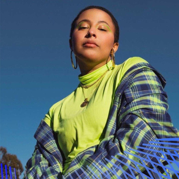 Walela has a shaved head, neon green eyeliner, and a slightly closed lipped smile. They wear a lime green turtleneck paired with a lime green and royal blue plaid blazer and pants and stand with their hands crossed near their abdomen.