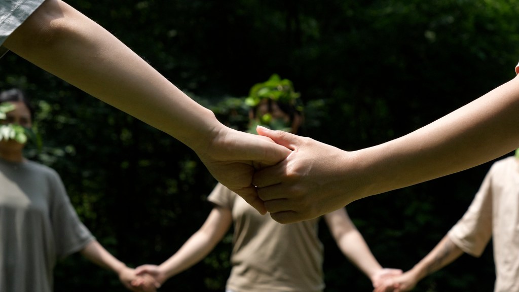 Close-up of two people holding hands in a circle, with others in the background, outdoors in a wooded area. The people are wearing pale earthy-tone t-shirts and leafy head pieces.