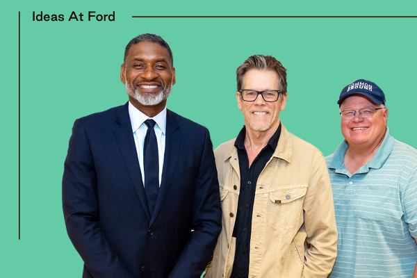 Three men stand side by side in front of a green background with “Ideas At Ford” written on it. Charles Blow, the first man on the left, wears a dark suit and tie. The middle man, Kevin Bacon, is wearing glasses and a beige jacket over a dark shirt. The man on the right, Gabe Brown, is wearing a blue cap and a striped polo shirt.