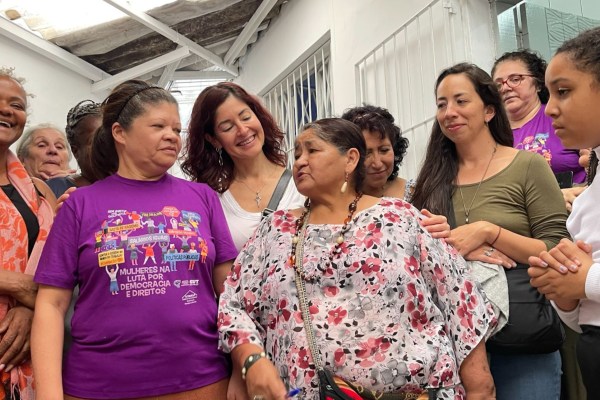 A cohort of LUNA graduates at the 2023 reunion in Brazil stands close together in a hallway. Some are smiling and engaged in conversation. One woman in front wears a floral blouse, while another wears a purple shirt with text and graphics.