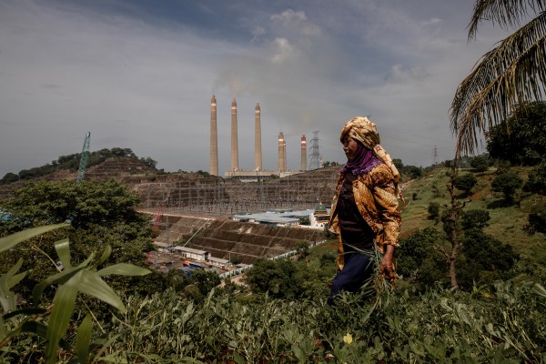 A person dressed in traditional clothing stands in a lush, green hillside overlooking a large construction site with multiple smokestacks in the background. The sky is partly cloudy, and there are various trees and plants around.