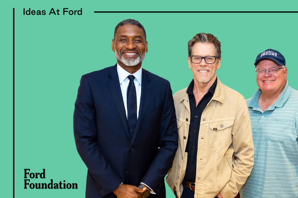 Three men stand side by side in front of a green background with “Ideas At Ford” and “Ford Foundation” written on it. Charles Blow, the first man on the left, wears a dark suit and tie. The middle man, Kevin Bacon, is wearing glasses and a beige jacket over a dark shirt. The man on the right, Gabe Brown, is wearing a blue cap and a striped polo shirt.