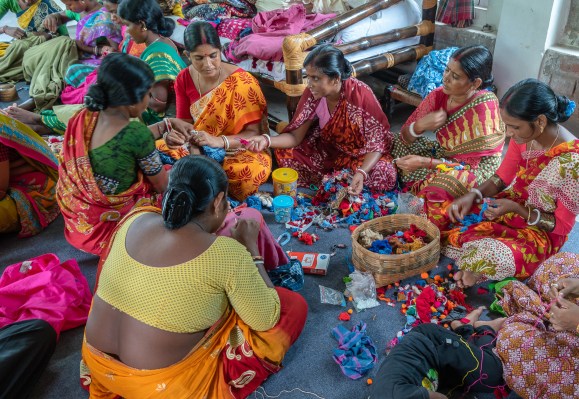 A group of women in colorful traditional attire sit on the floor, engaged in crafting activities. They work with various colorful threads and fabrics, surrounded by baskets filled with materials in a lively and collaborative setting.