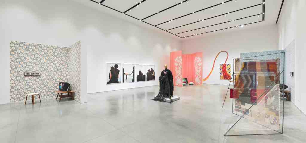 A white gallery interior with cement floors. There are multiple artworks in the gallery including a living room-inspired installation with a vintage television and floral wallpaper; a row of four photographs of women braiding their hair; a gold mannequin in a black dress, a pink curtain installation, a colorful canvas with paint.