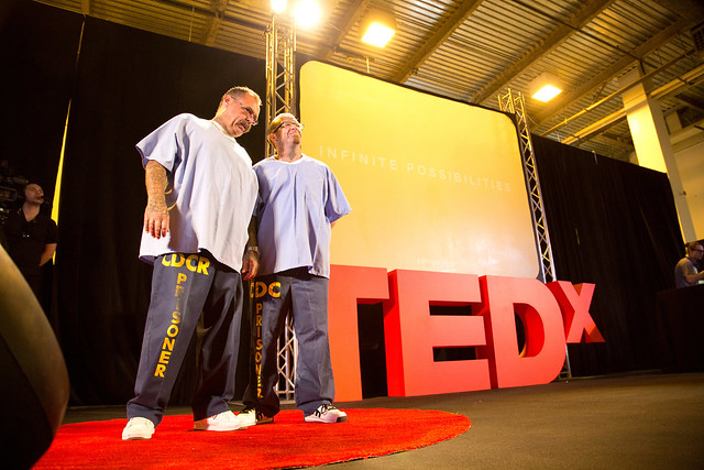 Two men in light blue shirts and dark pants with "PRISONER" written on them stand on a stage with a red TEDx sign. The background displays the words "INFINITE POSSIBILITIES." The setting appears to be a TEDx event.