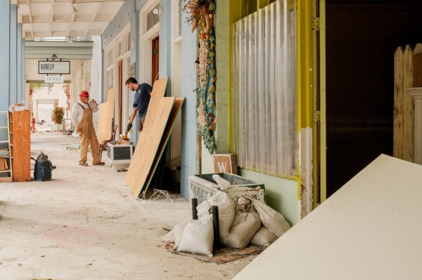 Two workers in a corridor are repairing, surrounded by wooden boards and tools. One is wearing overalls and a red cap, the other a blue shirt. The passage is lined with light blue walls and signs hanging above.