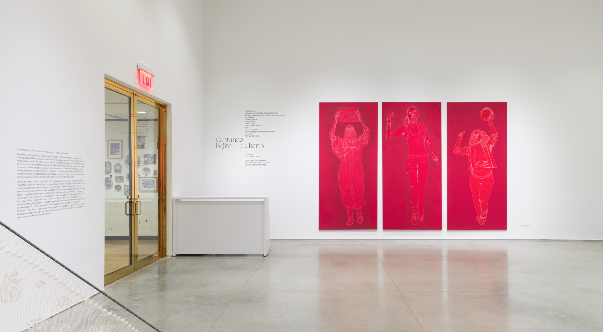 Gallery interior with white walls and a gray cement floor. A white desk is next to the entry doors. And to the right are three red tapestries featuring silhouettes of women protesters.