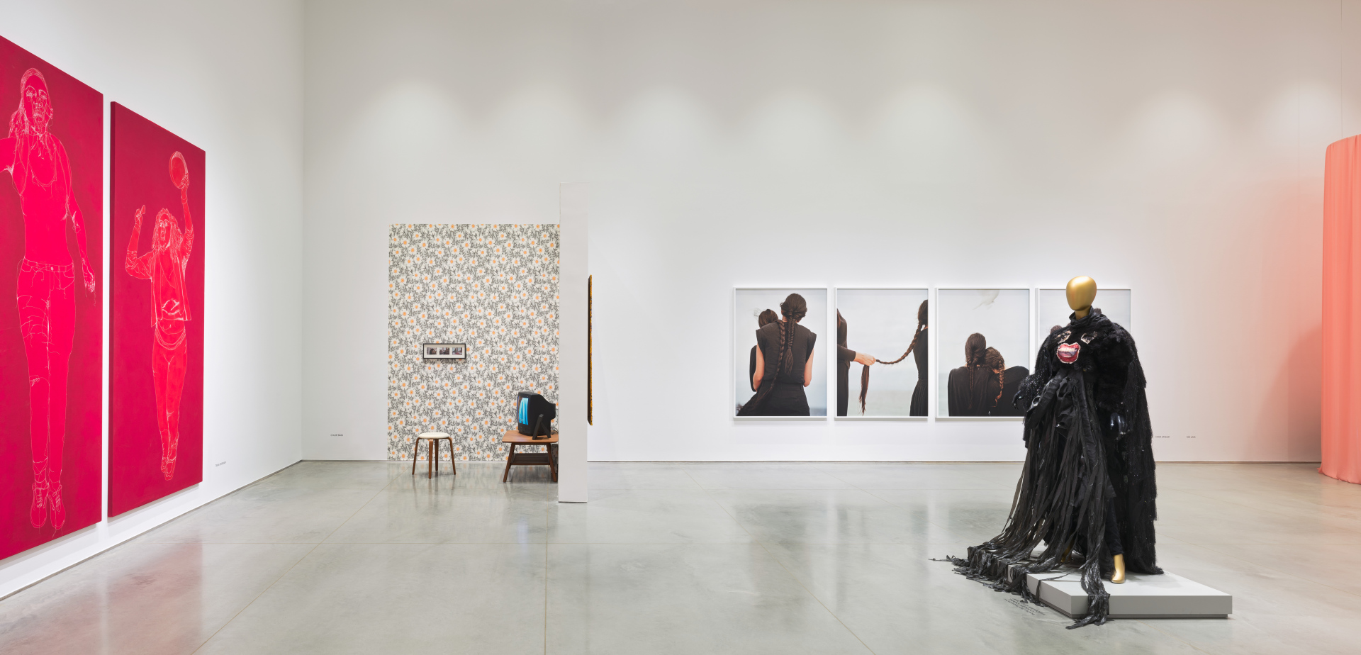 Wide-view of a gallery with a cement floor and multiple hanging artworks and installations including red tapestries; a living room installation; photographs of women braiding their hair; and a mannequin wearing a black dress.