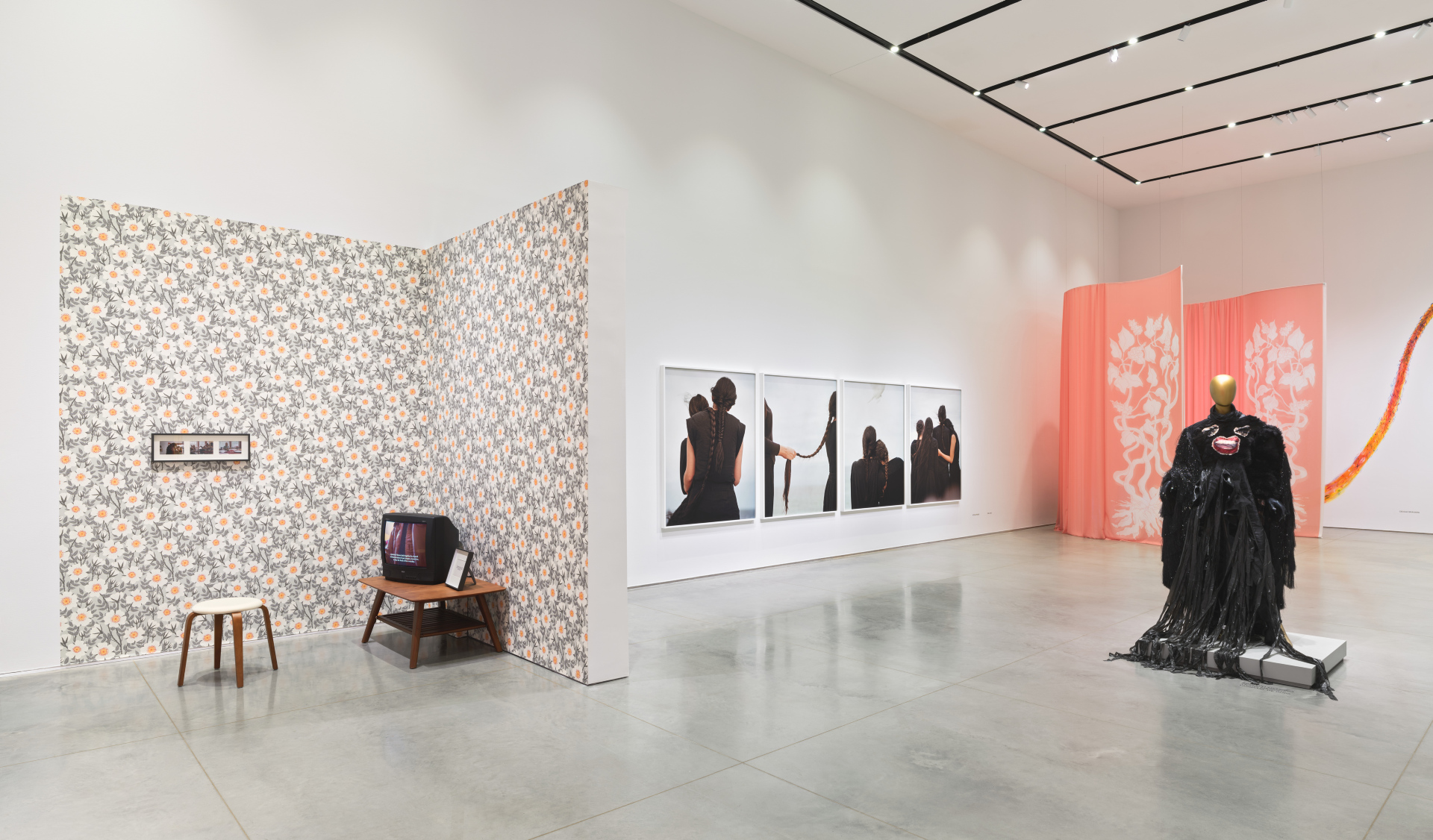 Gallery interior with multiple artworks and installations including a living room installation with floral wallpaper and a vintage television; photographs of women braiding their hair; a mannequin wearing a black dress, and a pink curtain.