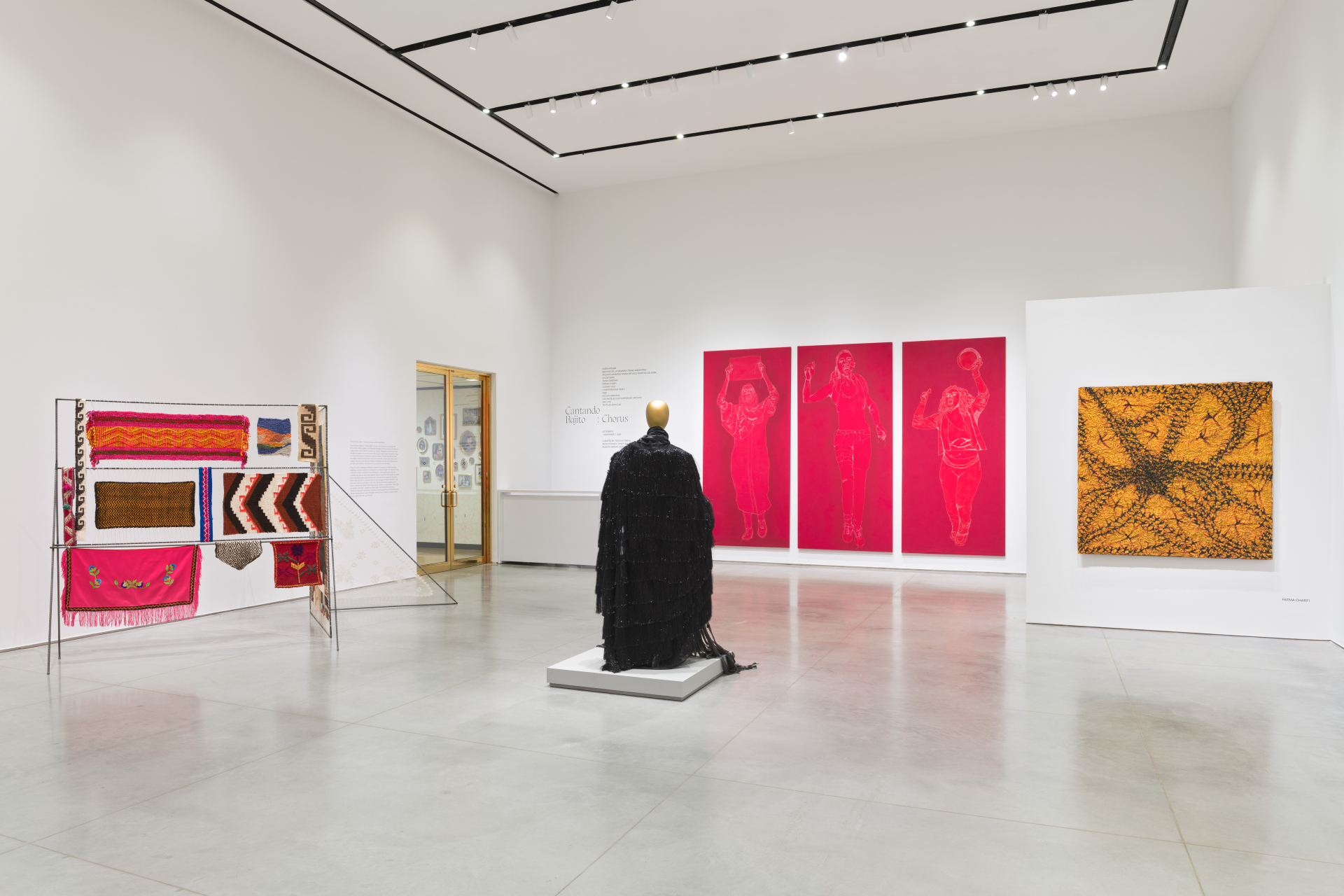 A gallery with multiple artworks and installations including a zigzag metal frame holding textiles; a mannequin wearing a black dress; red tapestries; and an abstract textured painting.