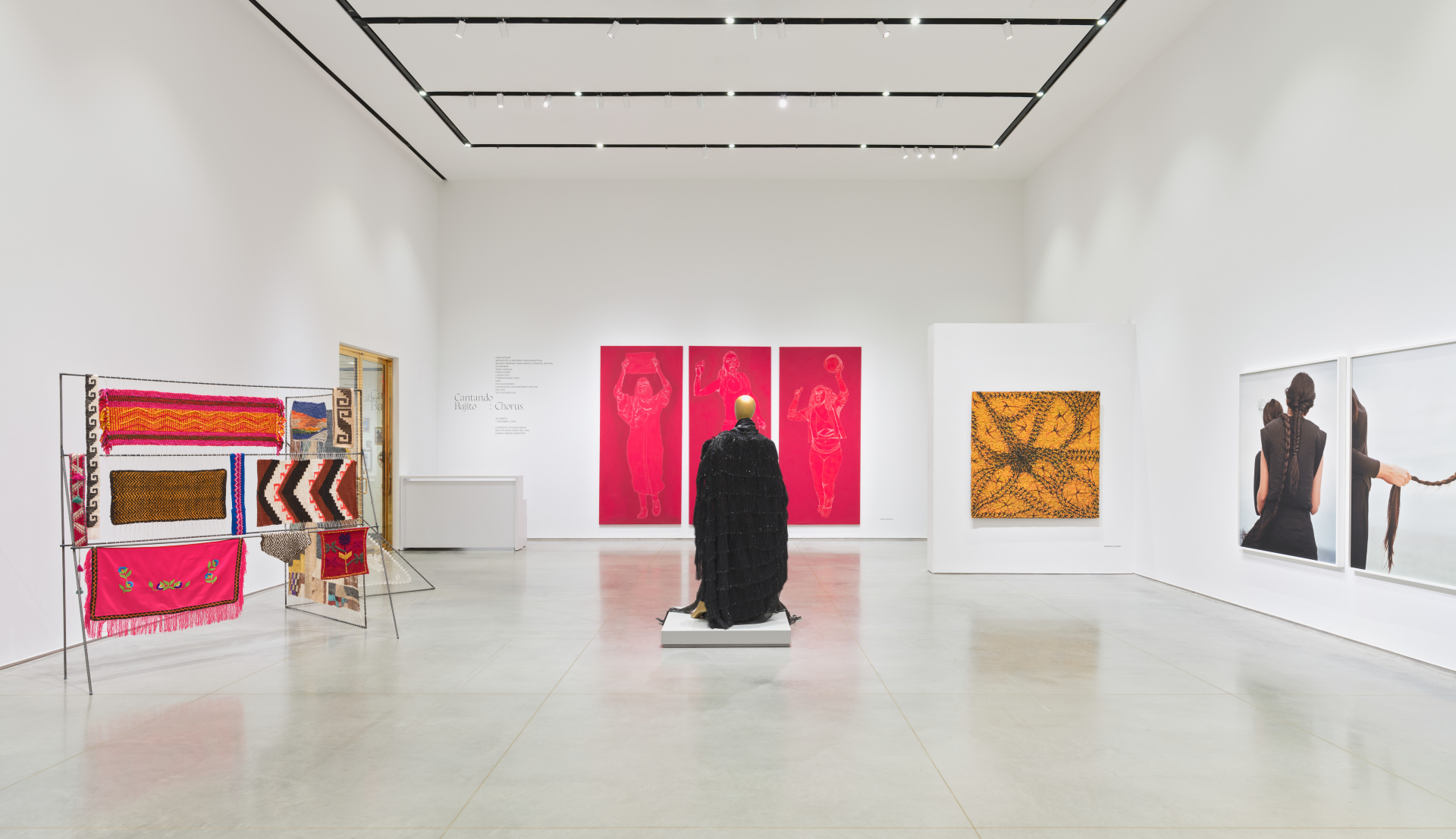 Gallery span of various artworks and installations including a zigzag metal frame holding textiles; a mannequin wearing a black dress; red tapestries; an abstract textured painting; and two photos of women braiding their hair.