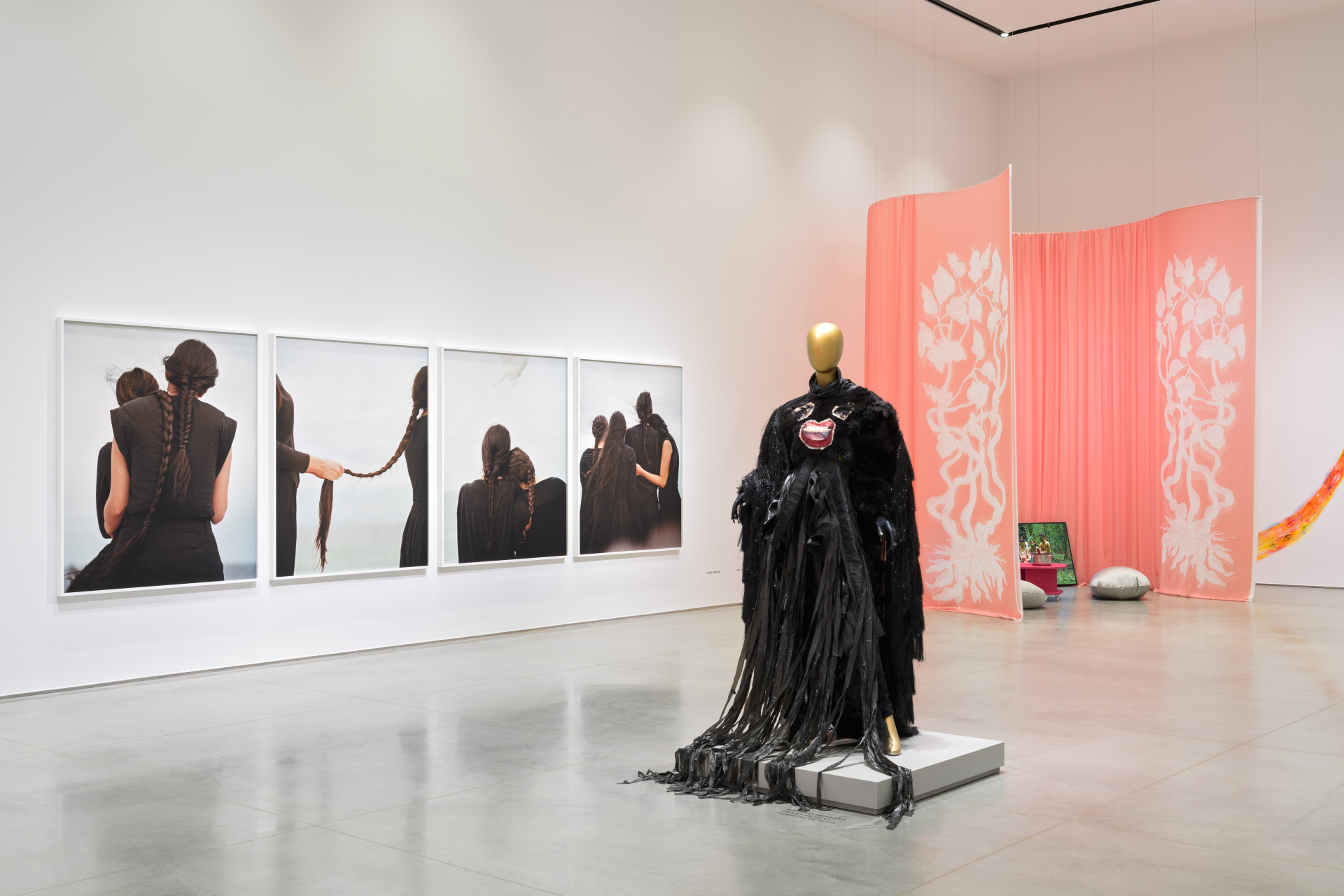 Gallery interior with four photographs of women braiding their hair; a mannequin wearing a black dress, and a pink curtain installation.