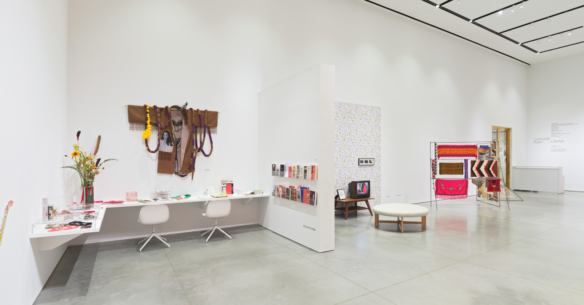 A gallery with a corner full of print materials and archival objects. And to the right, a vintage television installation against floral wallpaper, and a zigzag metal frame holding textiles.