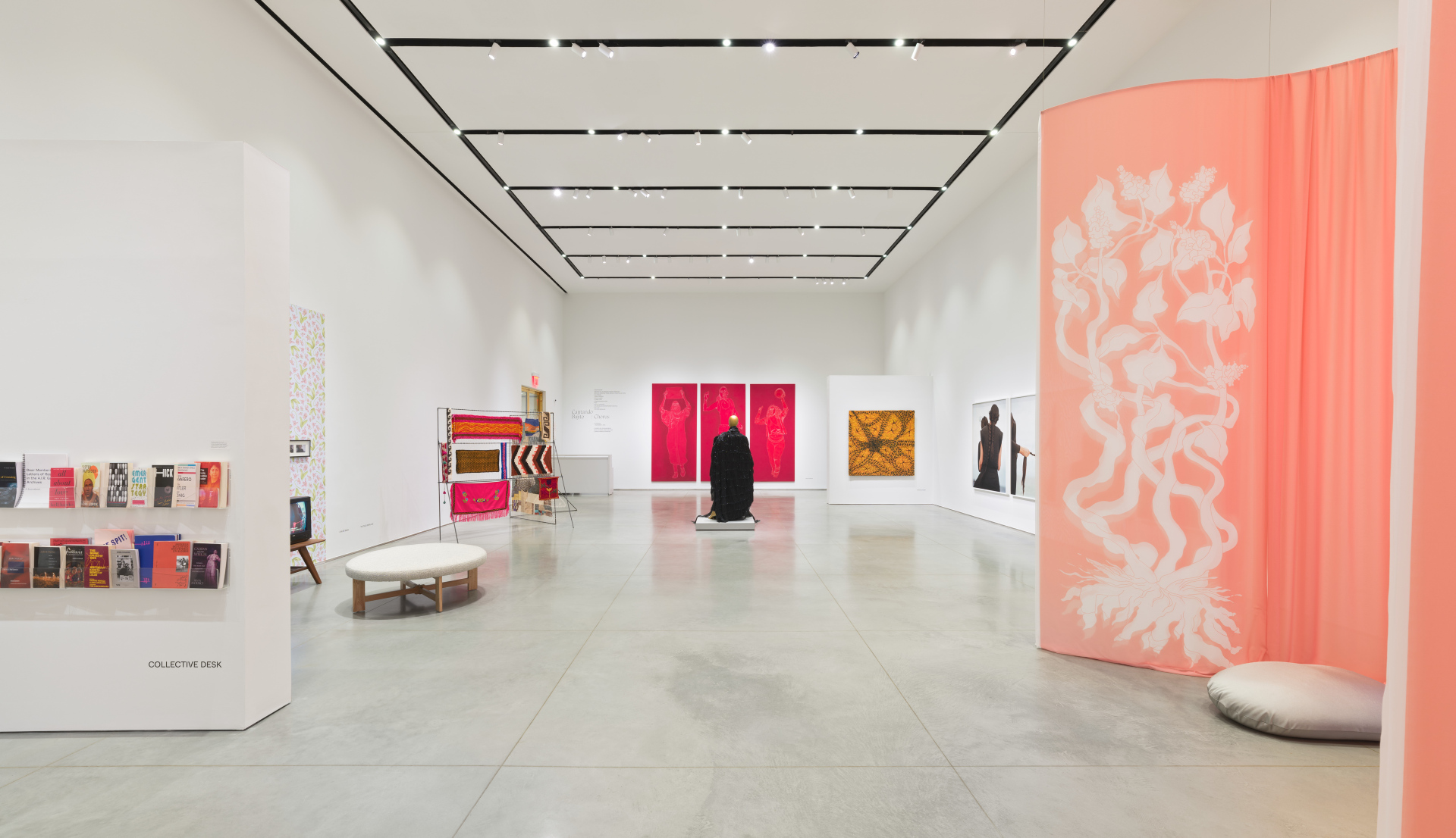 Gallery space with multiple installations, hanging artworks, rows of books, and cushions for seating.