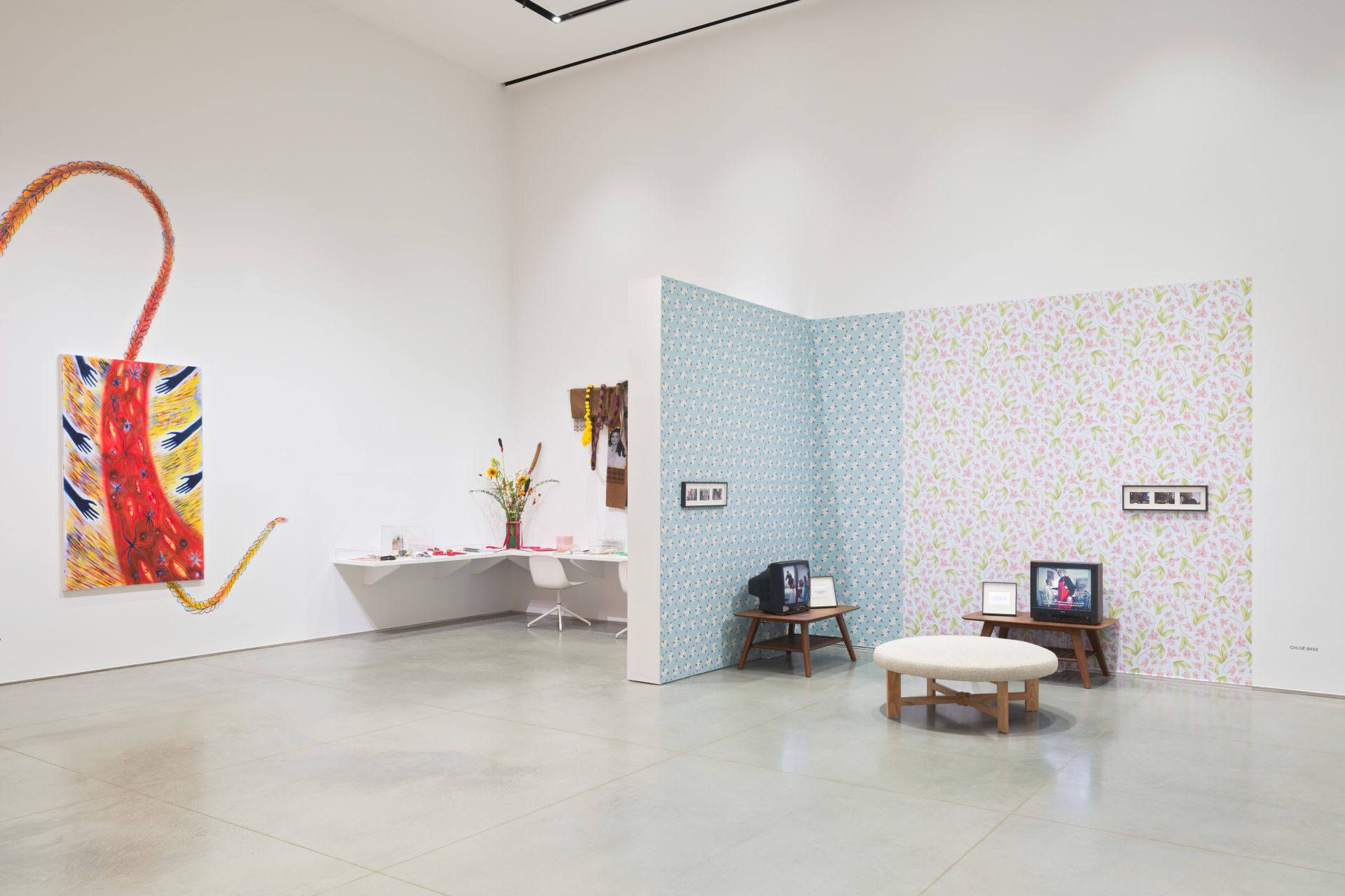 Corner of a gallery with an abstract wall painting, shelf scattered with archival objects, and a living room-esque installation with vintage televisions.