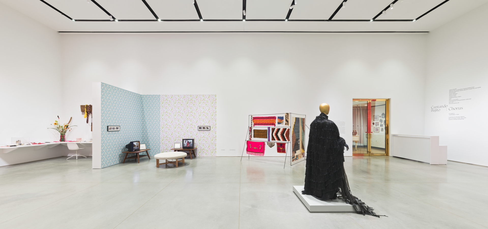 Wide view of a gallery with various artwork installations including a shelf of archival objects, television sets, textiles and a mannequin wearing a black costume.