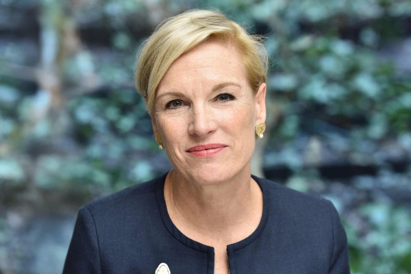 Cecile Richards with short blonde hair wearing a navy blazer and gold earrings, sitting in front of a blurred leafy background.