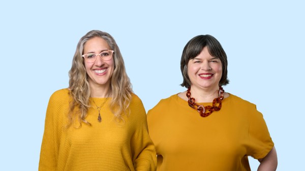 Two people are smiling in front of a light blue background. Both are wearing yellow tops, with one person having long, wavy hair and glasses, and the other having short hair and a bold necklace.